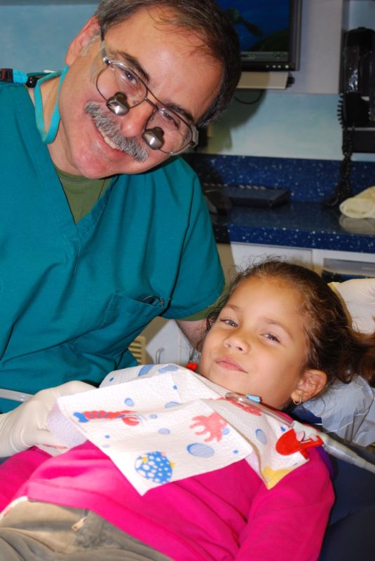 Dr. Joel Sokol leaning in and smiling next to a young girl resting in the treatment chair.
