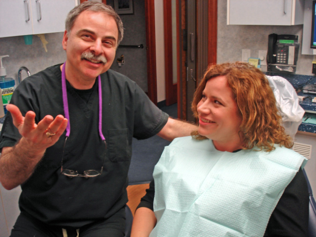 Dr. Joel Sokol in black surgical scrubs talking cheerfully with a patient seated in a dental chair.