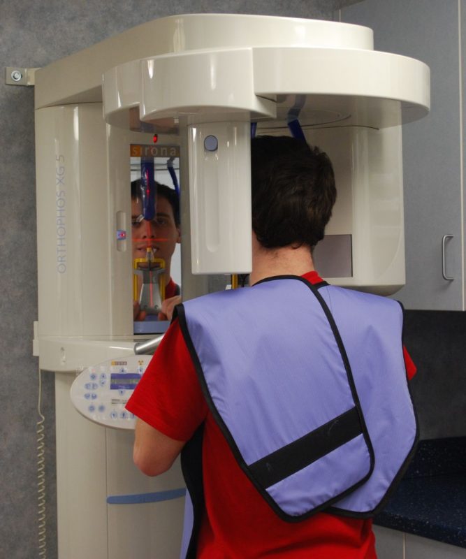Patient wearing a lead protective apron from behind while standing for a panoramic dental X-ray scan.