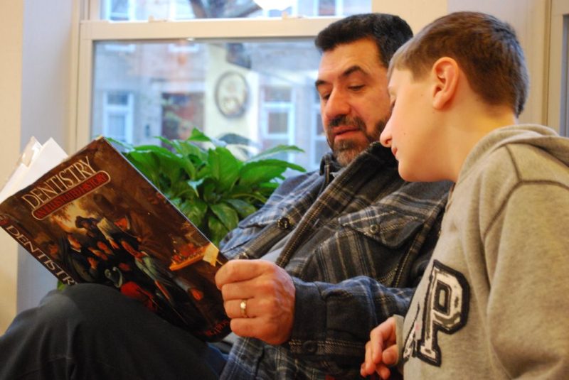 A man and a young boy sitting together in a waiting room, looking at an illustrated book about dentistry.